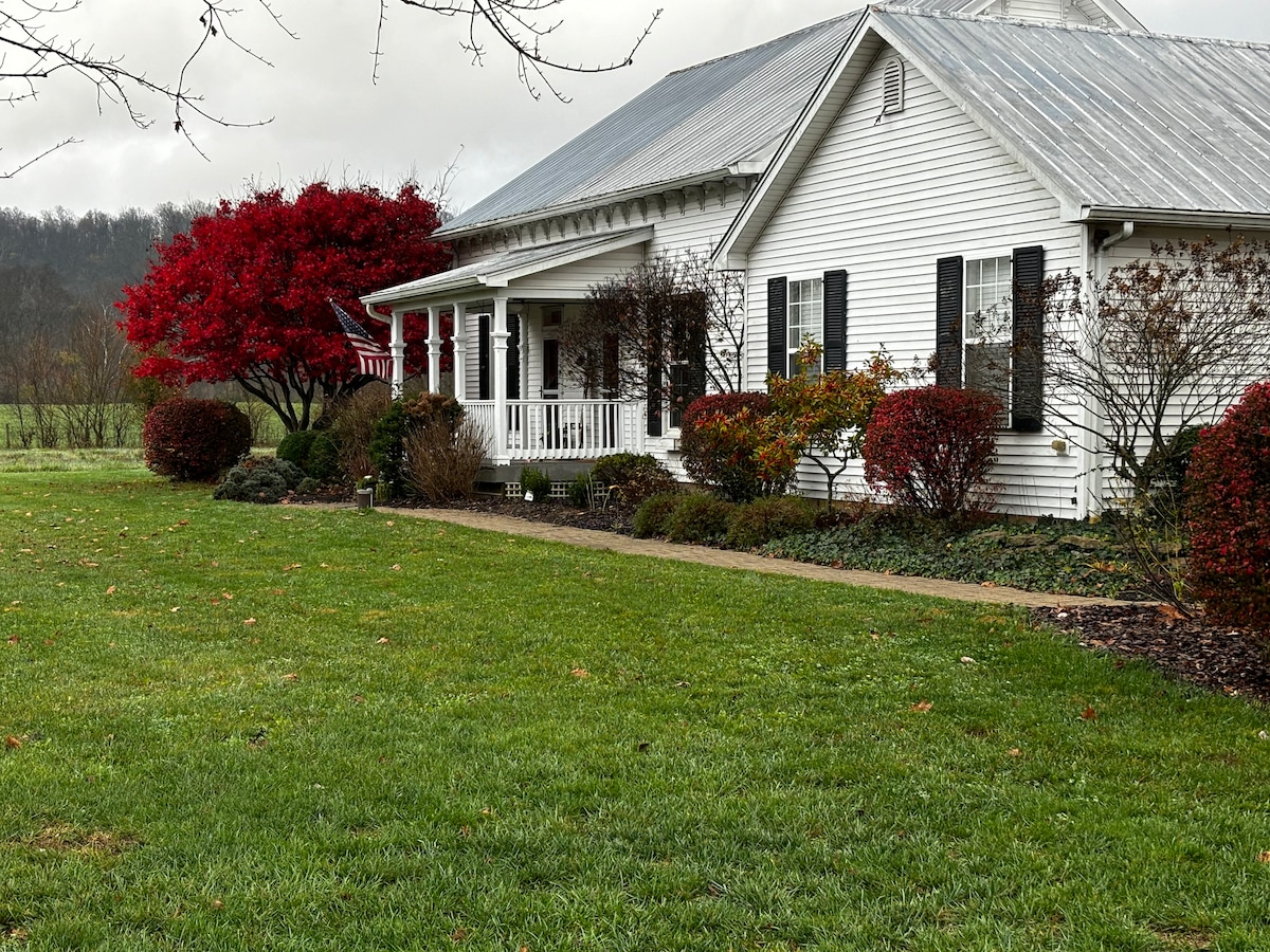 The exterior of the farmhouse is showcased, featuring a white wooden facade with a metal roof. Lush green grass surrounds the property, while colorful shrubs and a vibrant red tree add visual interest. A welcoming porch extends across the front, providing a space for relaxation.