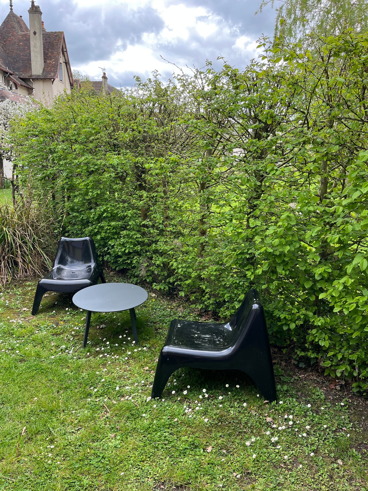 A small outdoor seating area is situated on a grassy patch, featuring two modern black chairs and a round table. Lush green foliage surrounds the space, with small white flowers dotting the ground, creating a private and serene atmosphere.