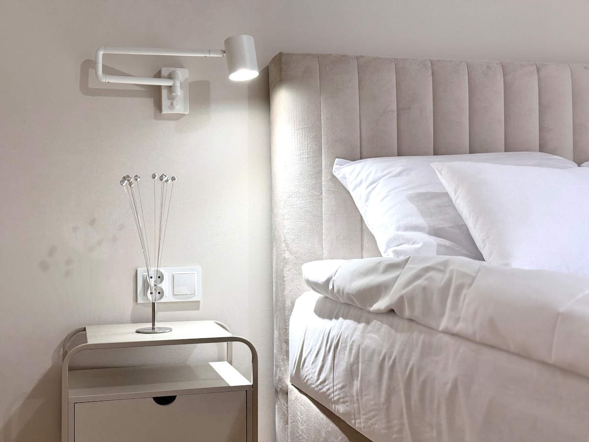 A serene sleeping area is presented with a spacious bed featuring a soft, tufted headboard. A bedside table holds a modern lamp and decorative elements. Soft linens and pillows create a calming ambiance, complemented by warm wall lighting.