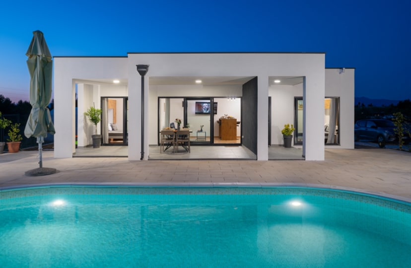 A modern white villa is highlighted under a twilight sky, featuring large glass doors that open to a spacious interior. A swimming pool reflects the night light, with lounge chairs surrounding the area and greenery enhancing the outdoor space.