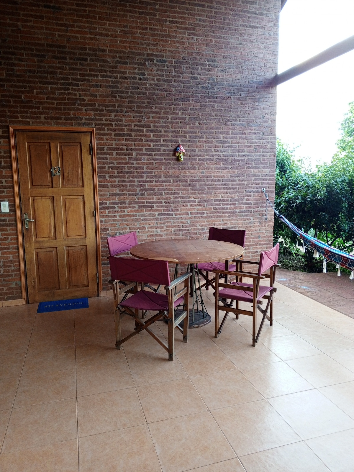 A terrace area is showcased with a round wooden table surrounded by four folding chairs. A wooden door leads inside, while natural light filters in from the nearby greenery, creating a relaxed environment. A hammock is visible in the background, adding to the outdoor appeal.