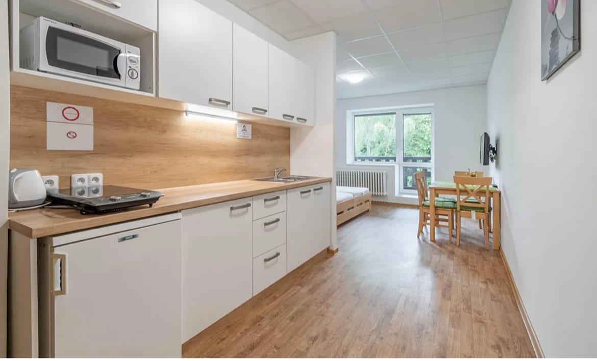 A clean kitchen area features modern cabinetry and appliances, including a microwave and refrigerator. A dining table for four is positioned by a window, allowing natural light to enter. A comfortable bed can be seen along the wall, enhancing the functional living space.