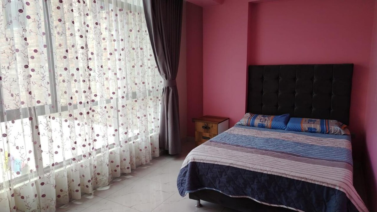 A bedroom features a spacious bed with a patterned blanket. Natural light filters through sheer curtains adorned with floral patterns, illuminating the room's pink walls. A bedside table is positioned next to the bed, providing additional space for personal items.