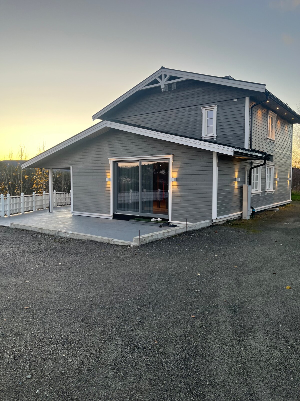The exterior of a two-story gray house is presented, featuring modern lines and a spacious deck. Large glass doors provide access to the interior, reflecting the evening light. A gravel driveway extends beside the home, bordered by a low fence and landscaped with tree views.