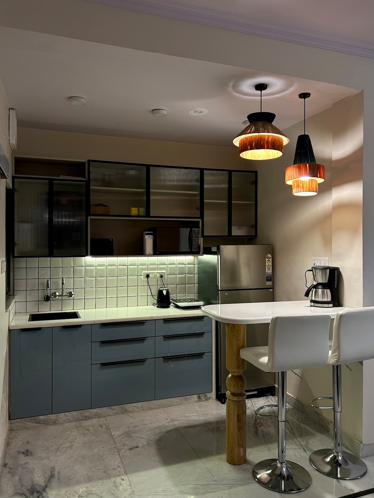 The kitchen features modern cabinetry with a chic design, complemented by a dining area with two stools. Task lighting illuminates the countertop, highlighting appliances such as a kettle, coffee maker, and refrigerator. Sleek marble flooring completes the contemporary look.
