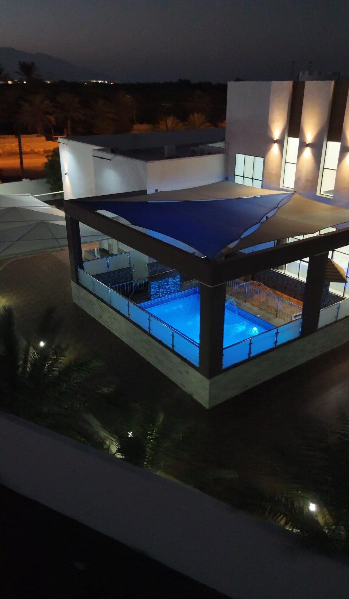 A private pool is highlighted under soft lighting, surrounded by a shaded seating area. The modern architectural lines of the structure complement the tranquil water, creating a serene outdoor setting. Surrounding palm trees are visible, adding to the peaceful ambiance.