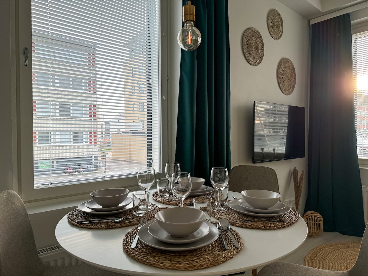 A round dining table is set with white dishware and glassware, ready for meals. Natural light filters through the blinds, highlighting the elegant seating and decorative wall plates. A television mounted on the wall is also visible.