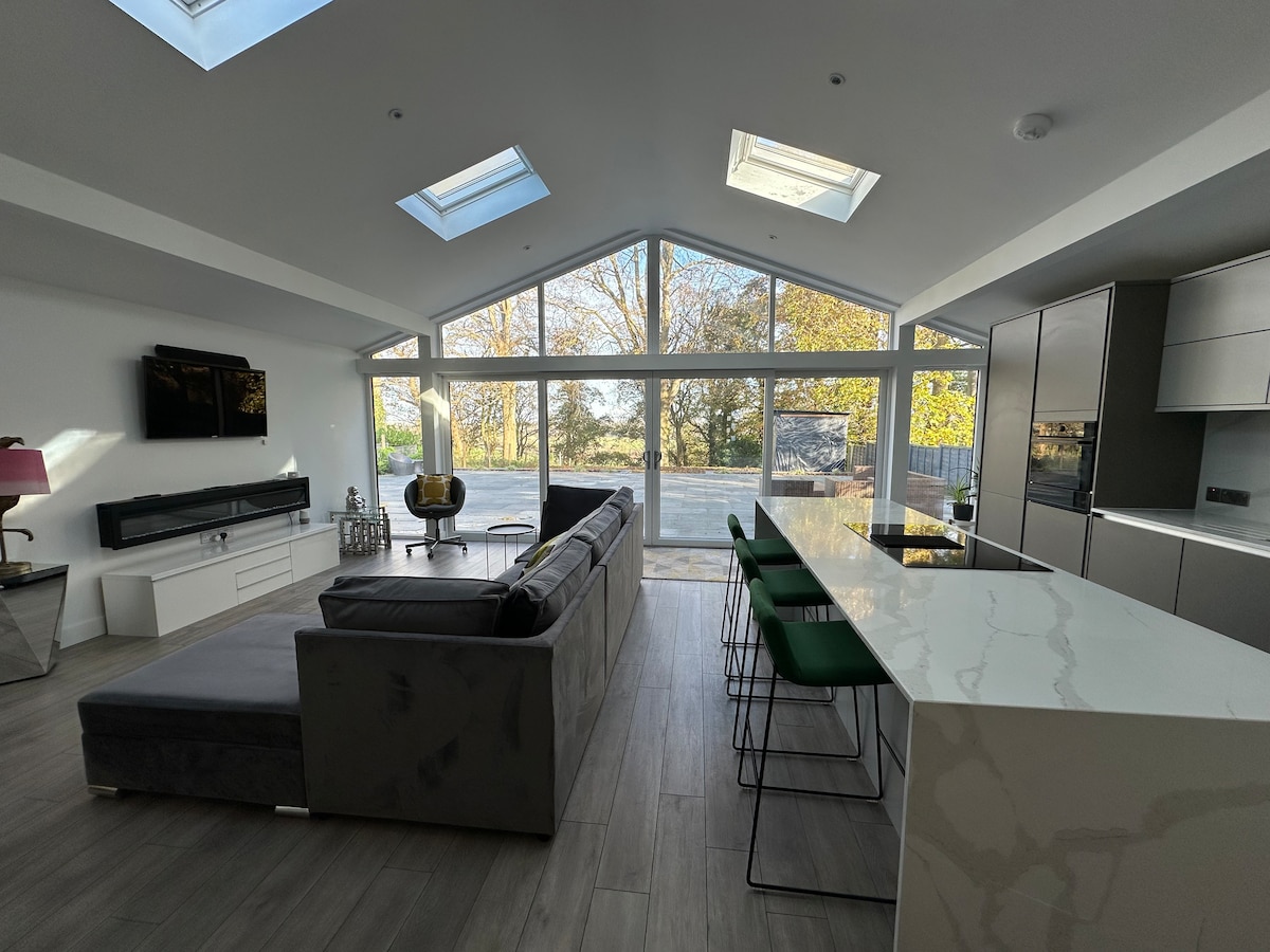 An open-plan living area features large glass windows, allowing natural light to flood the space. A comfortable gray couch faces a sleek entertainment unit, with a kitchen island in view, complemented by green bar stools. Skylights above enhance the airy ambiance.