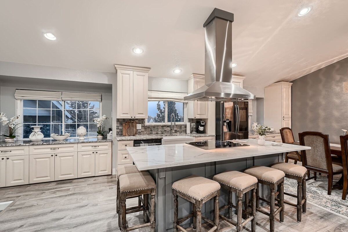 An open kitchen features a large central island with seating for six. Light cabinetry and marble countertops create a bright atmosphere, complemented by stainless steel appliances. Large windows provide natural light and a view of the outside, enhancing the spaciousness of the area.