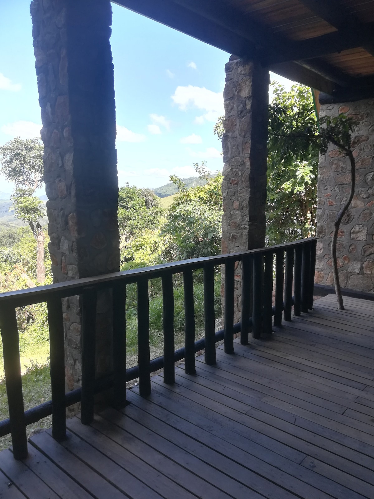 A covered patio features natural stone columns and wooden railings, providing an open view of the surrounding greenery. The wooden flooring adds a rustic touch, while the sky above is dotted with soft white clouds.