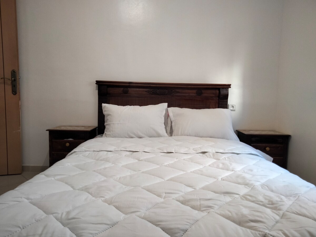 A cozy sleeping area is presented with a spacious bed featuring a white quilted blanket and two large pillows. Nightstands are placed on each side, contributing to a simple and functional layout. The walls are painted in a neutral tone, enhancing the room's brightness.