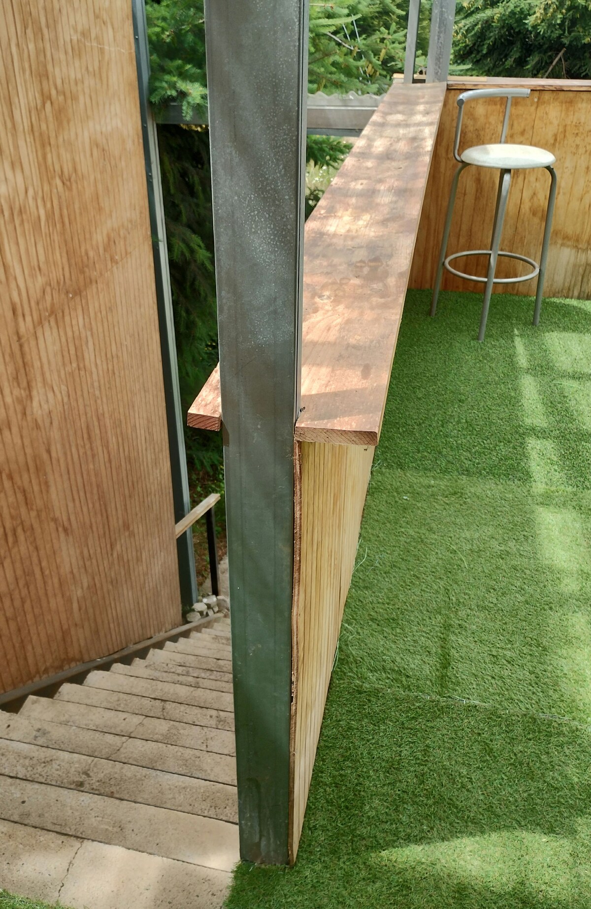 A staircase descends alongside a wooden wall, leading to a grassy outdoor area. A bar-like counter is visible, featuring a narrow copper tabletop and a metal stool. Soft green grass covers the ground, creating a pleasant and natural environment.