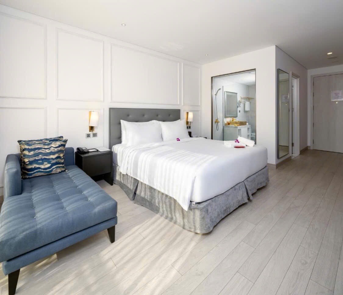 A spacious room features a large bed with crisp white linens and decorative cushions. A comfortable blue lounge chair is positioned beside the bed. Natural light fills the space through a large window, with a separate bathroom visible through glass doors.