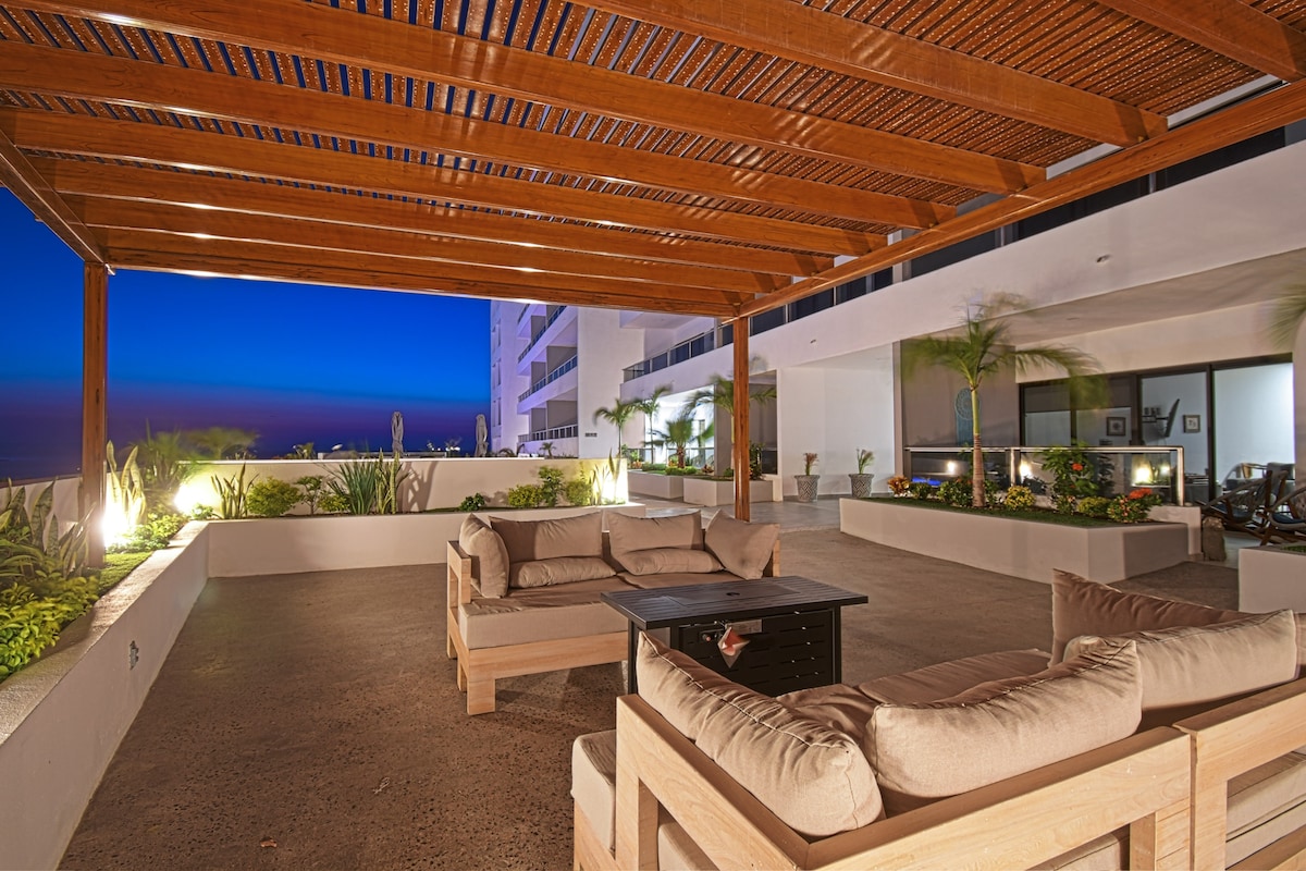 A spacious outdoor seating area is illuminated under a wooden pergola, featuring comfortable sofas arranged around a central table. Lush greenery lines the perimeter, and ambient lighting enhances the tranquil atmosphere during twilight.