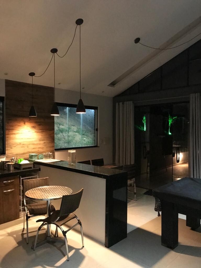 The kitchen area features modern fixtures with a sleek black countertop and wooden cabinetry. A round table with four chairs is positioned nearby. Large windows illuminate the space, providing views of the surrounding nature, while pendant lights add a touch of ambiance.