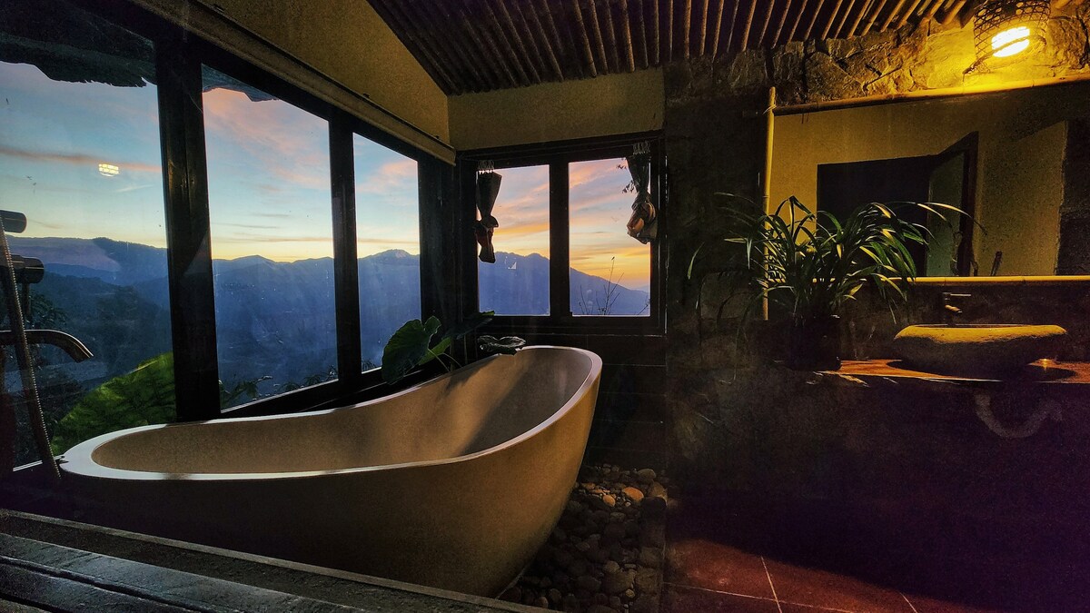 A spacious bathroom features a freestanding bathtub positioned near large windows that offer expansive views of the surrounding mountains. Natural light from the sunset casts warm tones throughout the space, highlighting the stone accents and greenery near the bathtub.