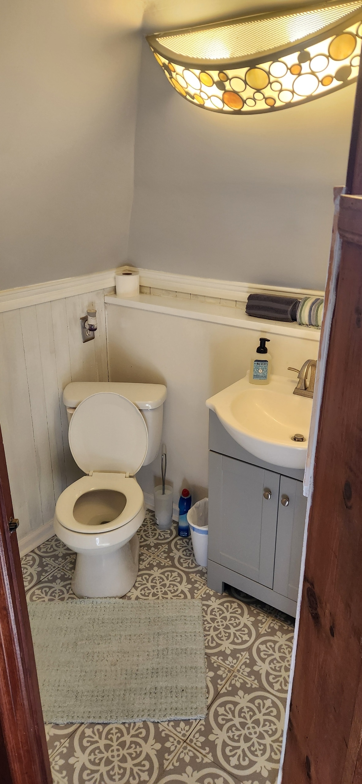 A compact bathroom features a modern sink with a light-colored cabinet and a round mirror above it. A white toilet is positioned beside the sink. The space is adorned with patterned flooring and decorative wall trim, with soft lighting provided by an overhead fixture.