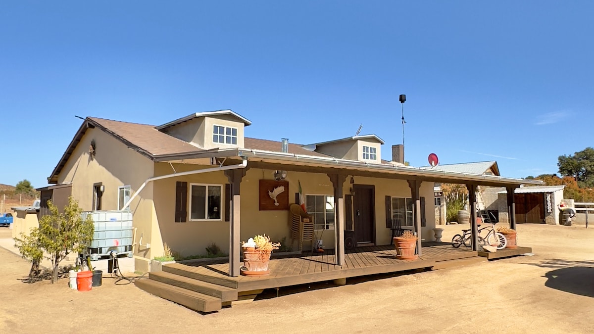 The home's exterior features a covered porch with wooden beams, adorned with pots of flowers. A light-colored facade and multiple windows provide natural light, while a gravel driveway leads to additional structures in the background. The clear blue sky adds to the inviting atmosphere.