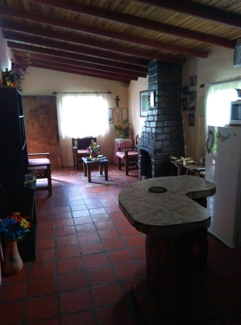 The cozy interior features terracotta tile flooring and wooden beams. A rustic fireplace is situated against a stone wall. A small table is positioned in the foreground, with wooden chairs arranged around it. Natural light enters through windows adorned with light curtains.