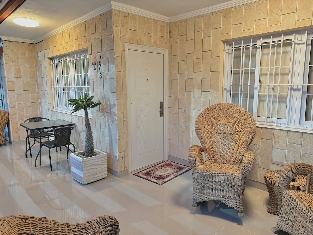 A welcoming entryway is presented with light-toned decorative wall tiles. Two woven chairs are positioned near a small glass-top table, creating a comfortable seating area. A potted plant adds a touch of greenery, while the door features a simple design and modern hardware.