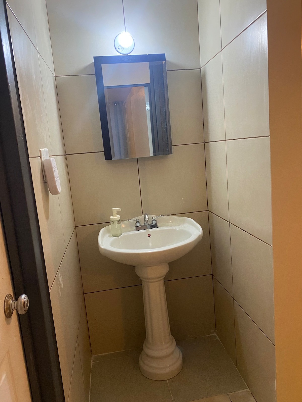 A compact bathroom features a pedestal sink with chrome fixtures, positioned beneath a wall-mounted mirror. Soft lighting above the mirror provides illumination, while taupe tiles cover the walls and floor, creating a clean and functional space.