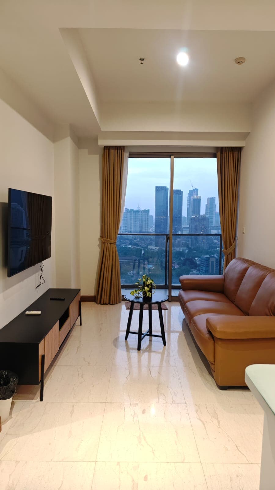 A living area features a brown leather sofa facing a wall-mounted television. A small round table is positioned beside the sofa, adorned with a simple floral arrangement. Natural light filters in through a large window, offering views of the city skyline beyond.