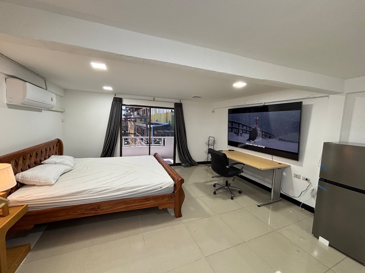 A spacious room contains a wooden bed with a plain mattress and a desk facing a large screen. Natural light enters through a wide window, covered by dark curtains. A refrigerator is positioned nearby, and air conditioning is visible on the ceiling.