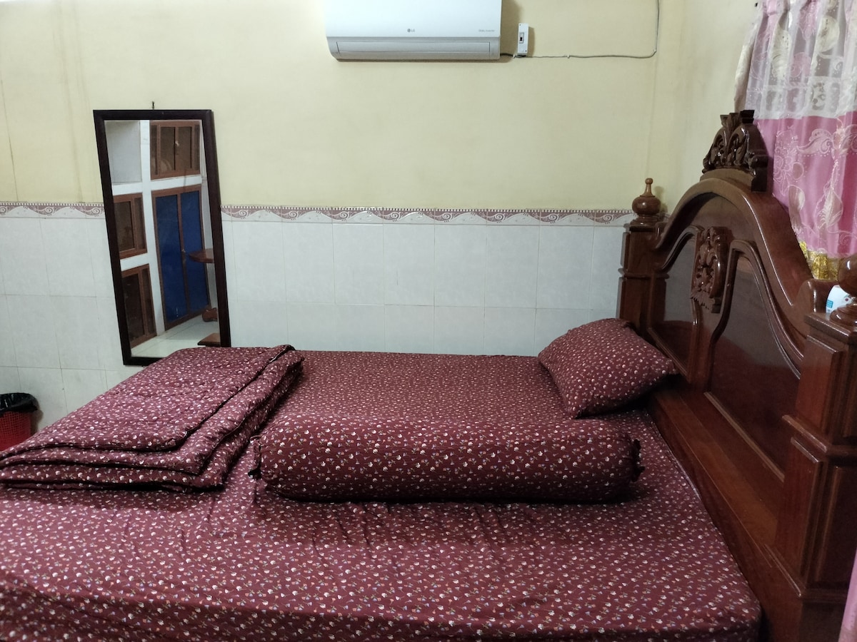 The sleeping area features a wooden bed with burgundy patterned bedding. A matching bolster and pillows are neatly arranged, while a wall-mounted air conditioner is visible above. A mirror reflects the room, complementing the light-colored walls and tiled floor.