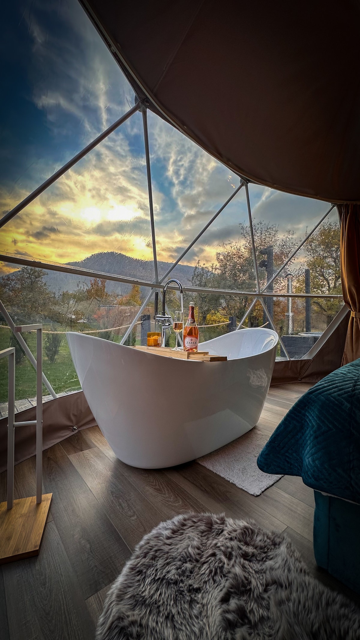 A freestanding bathtub is positioned by large panoramic windows, offering views of the natural landscape outside. A wooden tray holds drinks beside the tub, while soft textures are provided by a cozy area rug on the floor. Gentle lighting enhances the serene environment.
