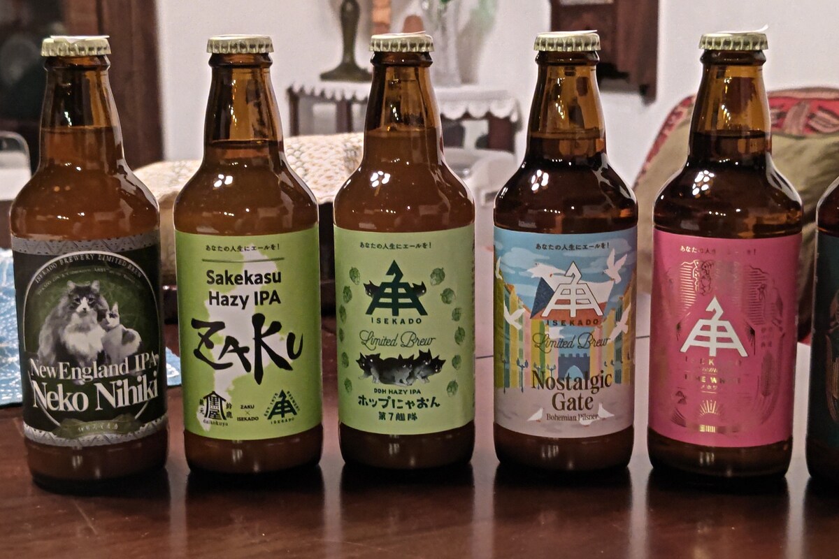 Five bottles of craft beer are arranged on a wooden table. Each bottle features distinct labels with colorful designs and typography. Varieties include New England IPA, Sakekasu Hazy IPA, Rhiador Bear, Nostalgic Gate, and others, highlighting a variety of flavors and styles.