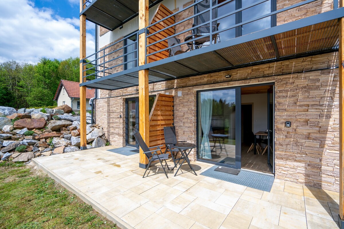 A spacious patio area is shown, featuring a small table and two chairs positioned on a tiled surface. Natural stone walls are complemented by wooden accents. Large glass doors provide access to the interior, with a view of greenery in the background.