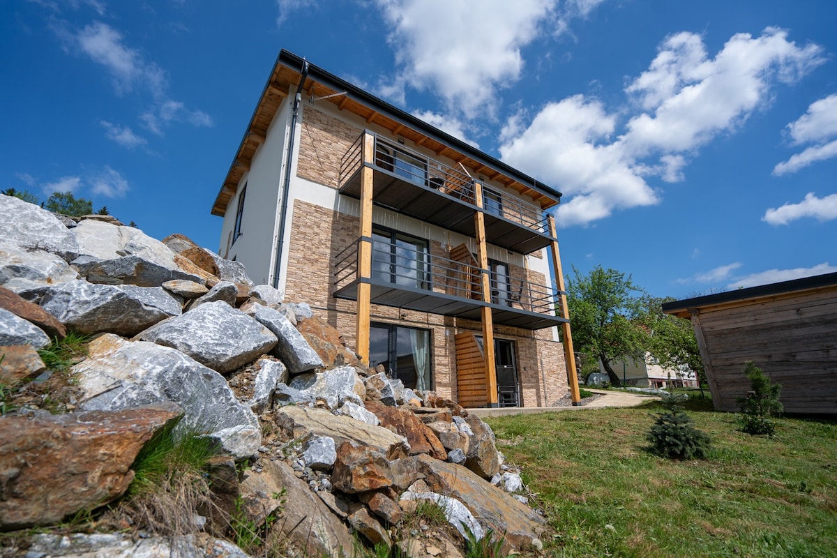 A modern building is showcased from an outdoor angle, featuring a combination of stone and wooden elements. The structure has multiple balconies with railing, set against a clear blue sky and surrounded by greenery and rock formations.
