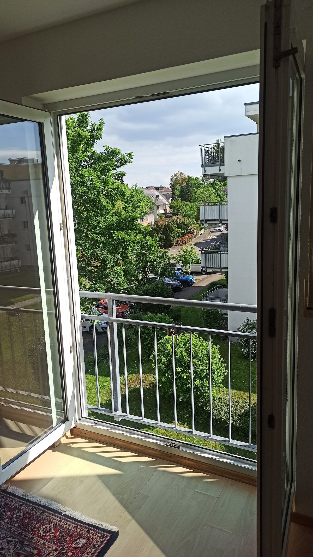 A balcony door stands open, revealing views of lush greenery and nearby residential buildings. The spacious outdoor area is framed by a simple railing, while light streams into the room from the clear glass doors. A rug is visible on the floor inside.