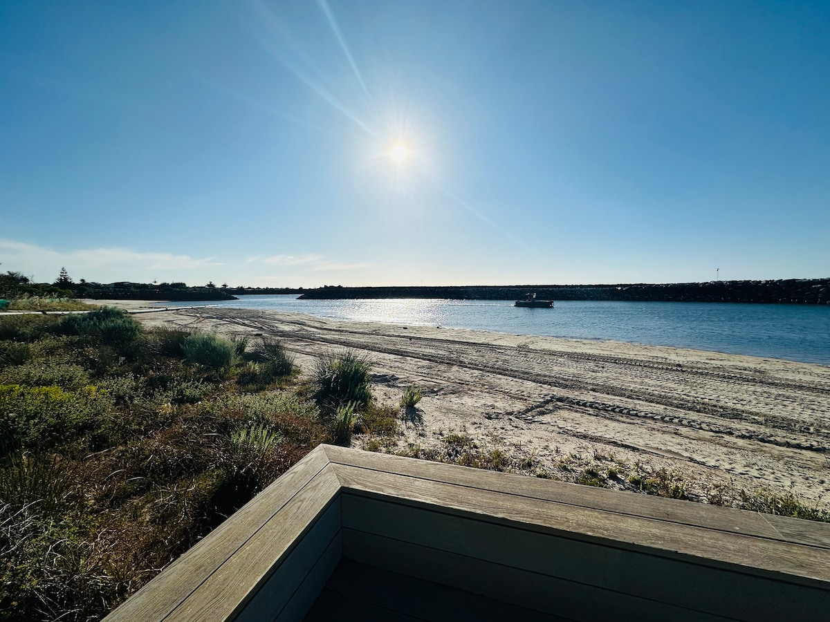 A serene beach view is captured, featuring soft sand and gentle waves lapping at the shoreline. Bright sunlight reflects off the water, while footprints in the sand and a distant boat suggest leisure activities. Coastal vegetation borders the area, enhancing the tranquil setting.
