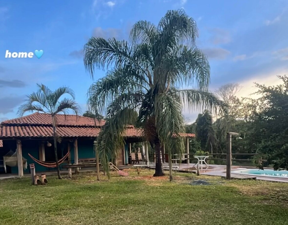 A spacious outdoor area is shown, featuring a well-maintained garden with palm trees and a hammock. A rustic building with a tiled roof is visible in the background, alongside a wooden deck and pool. Soft lighting is created by the setting sun.
