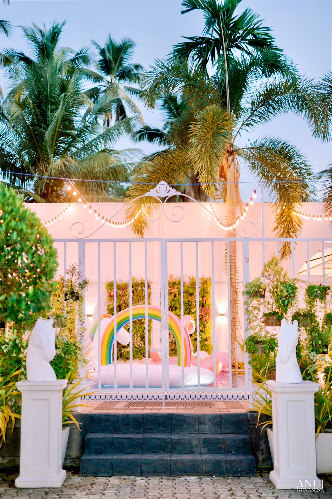 A welcoming entrance is framed by lush greenery and palm trees, with soft lighting illuminating the area. A decorative gate stands invitingly open, revealing a vibrant rainbow and a cozy seating arrangement within the serene garden.