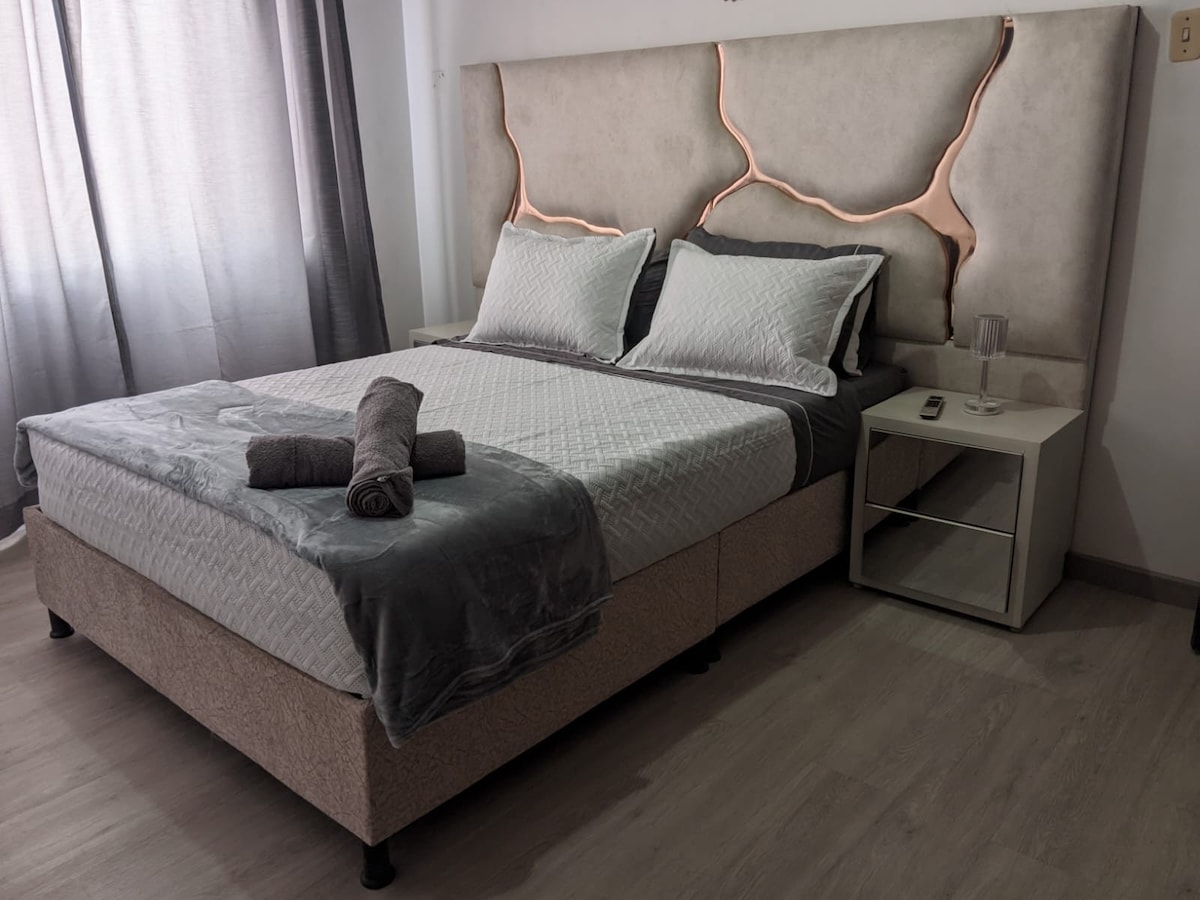 A spacious bed is framed by a modern headboard with elegant detailing. Soft grey linens and rolled towels are neatly placed atop the bed. A bedside table holds a glass and a small remote, while natural light enters through sheer curtains.