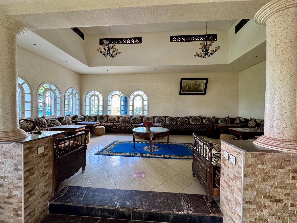 A spacious living area features multiple large windows that allow natural light to fill the room. Comfortable seating is arranged along the curved walls, complemented by decorative cushions. A central table sits on an ornate rug, with elegant chandeliers hanging above, enhancing the inviting ambiance.
