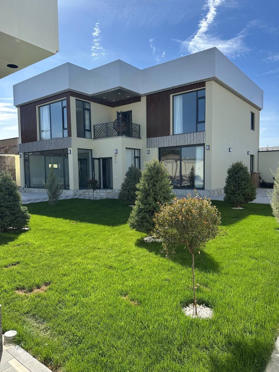 A modern two-story house is surrounded by lush greenery, featuring large windows that invite natural light. A well-maintained lawn with small shrubs and trees enhances the exterior. The facade combines contemporary lines with wooden accents, creating a harmonious appearance.