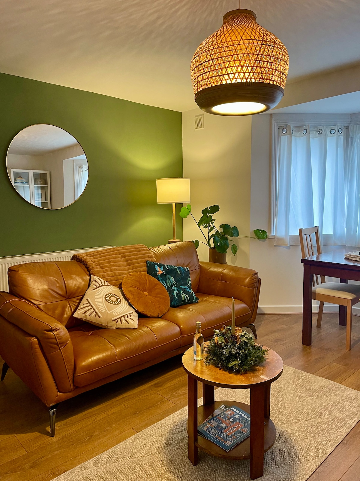 A comfortable lounge area features a leather sofa adorned with decorative cushions. A round mirror reflects soft lighting from a standing lamp. A wooden table is positioned beside a small coffee table, while large windows with light curtains allow natural light to fill the space.