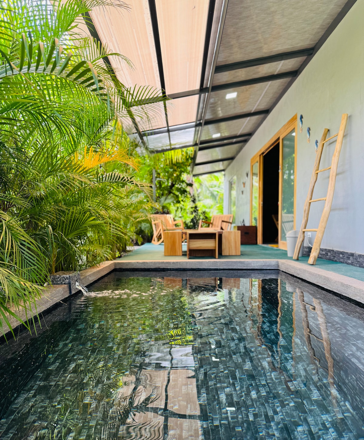 A tranquil outdoor area features a small water pool reflecting light and surrounded by lush greenery. Comfortable seating areas are positioned nearby, offering a space for relaxation. The entrance to the interior is visible in the background, framed by the open structure.