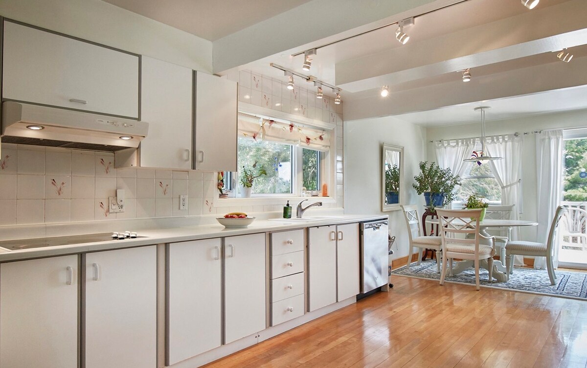 A bright kitchen features white cabinetry and a large window overlooking a dining area. Modern appliances, including a dishwasher, are present. Light hardwood flooring extends throughout, and a small dining table with four chairs is positioned near the window, complemented by tasteful decor and natural light.