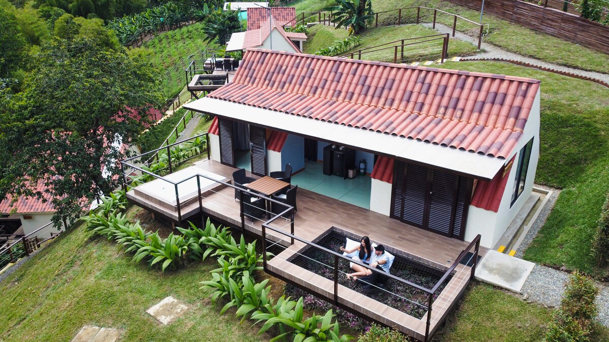 The exterior of a charming rural accommodation is showcased, featuring a terracotta-tiled roof and a wooden deck. A small plunge pool is surrounded by greenery, while lounge chairs invite relaxation. Scenic hills create a peaceful backdrop, enhancing the outdoor living space.