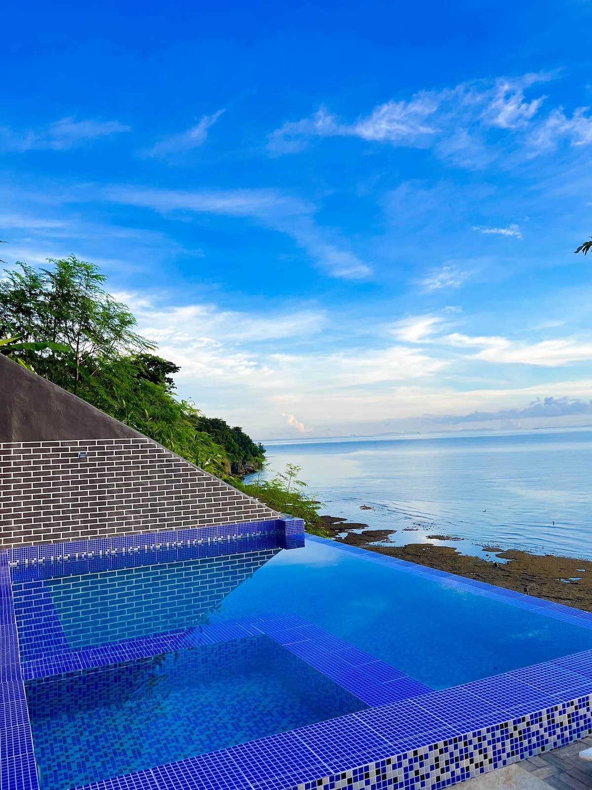 An infinity pool stretches towards the horizon, featuring mosaic tiles in shades of blue. Lush greenery lines the edge, contrasting with the expansive sky above. The calm water reflects the serene landscape, inviting relaxation and tranquility by the seaside.