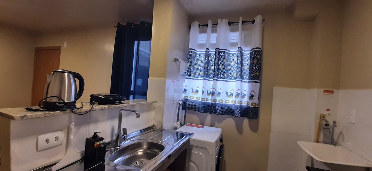 A well-equipped kitchen is present, featuring a stainless steel sink and a washing machine. Natural light enters through a window adorned with a patterned curtain. A kettle and other kitchen essentials are visible on the countertop, contributing to a functional cooking space.