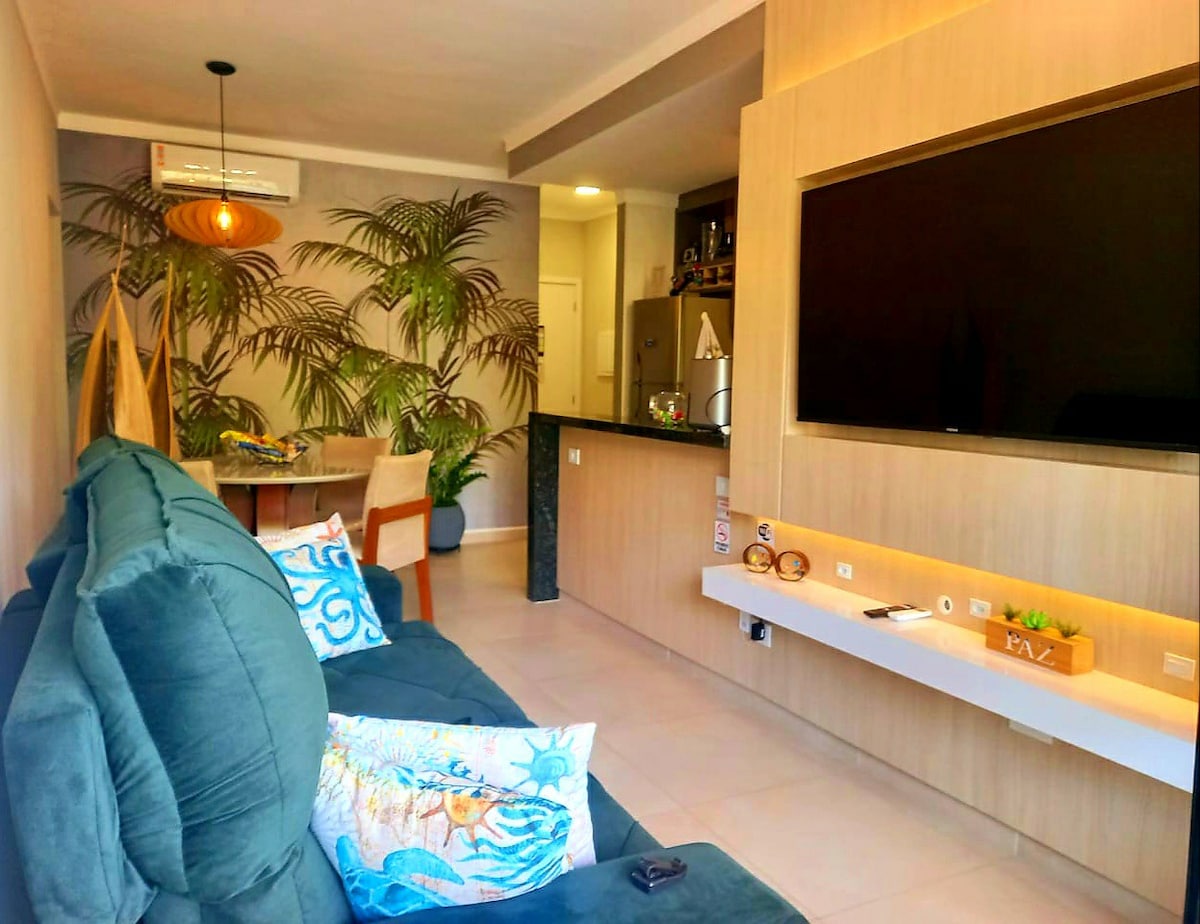 A modern living area features a large green sofa adorned with decorative pillows. A flat-screen TV is mounted on the wall above a light-colored console. The space is complemented by indoor plants and a dining table in the background, creating a bright and inviting environment.