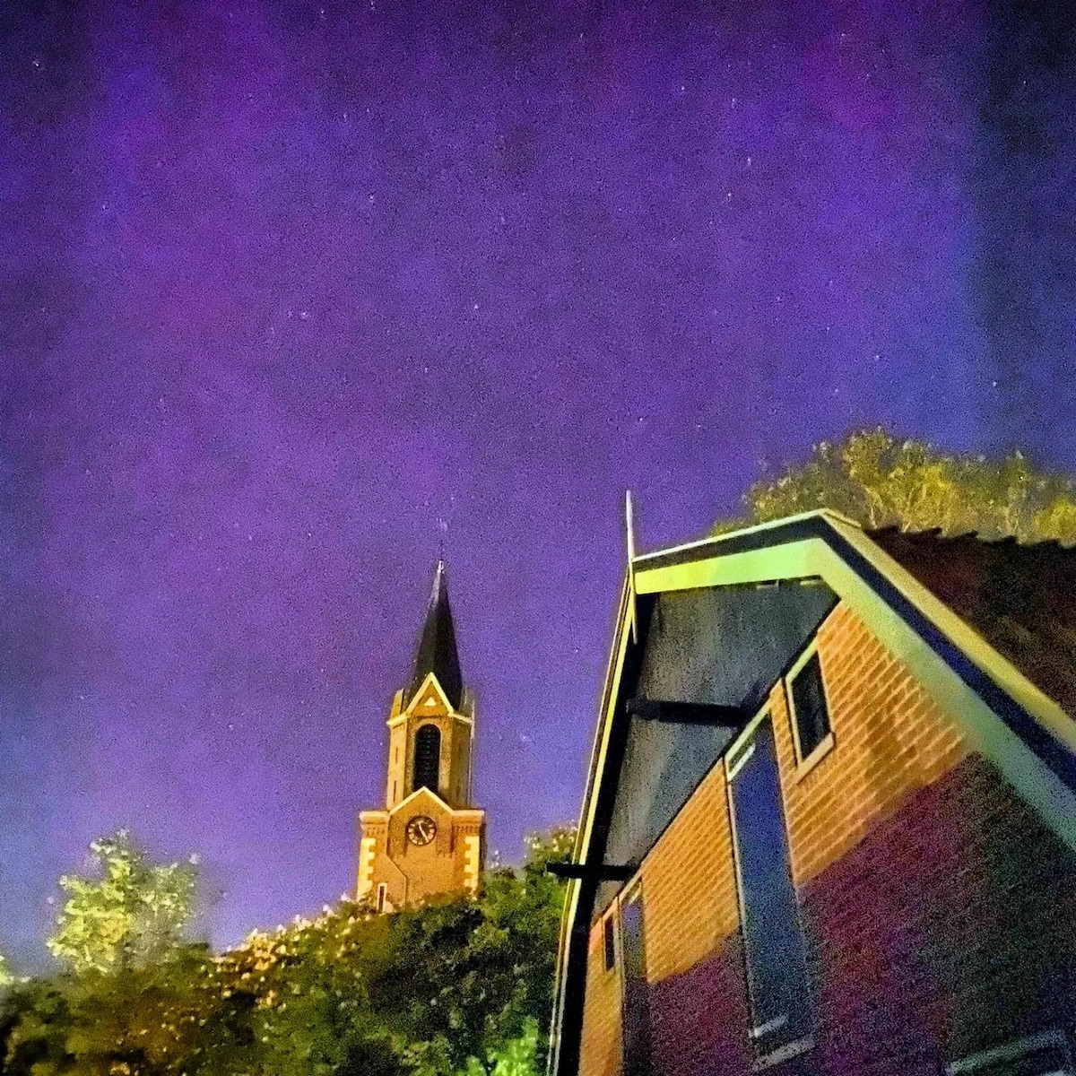 A nighttime view captures the historic church tower rising against a dark purple sky, with stars visible above. An angled roof of a nearby house adds to the composition, highlighting the serene ambiance of the setting.