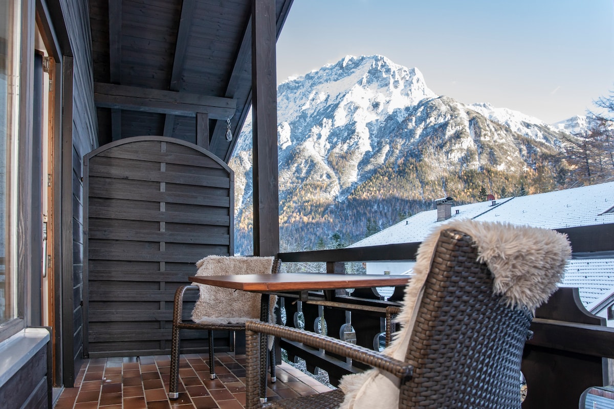 A balcony is shown with a wooden railing and a table accompanied by two chairs. Textured cushions add comfort. A stunning mountain view is visible in the background, with snow-capped peaks and hints of autumn foliage. The setting offers a serene outdoor space.