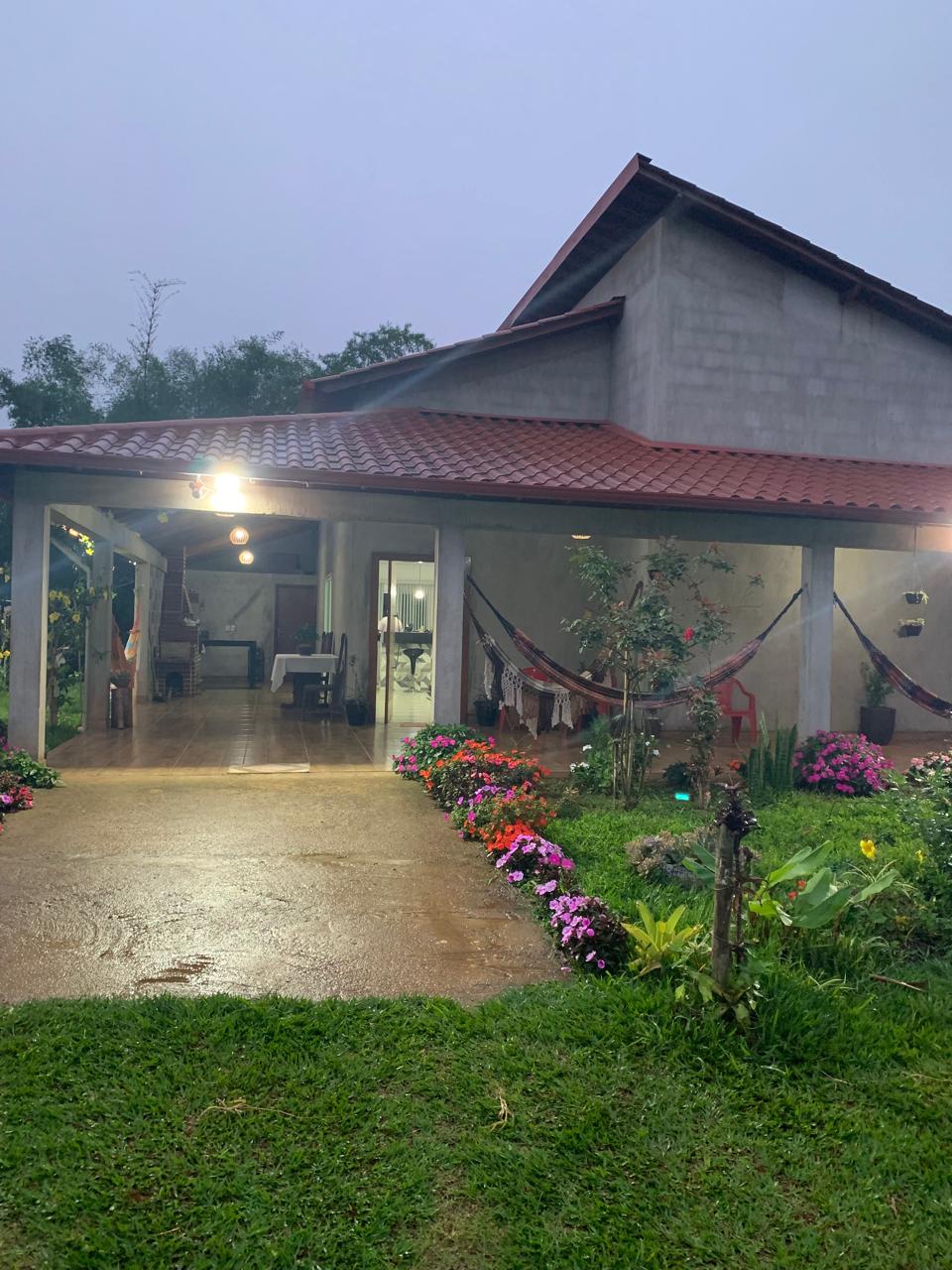 The exterior of a spacious home is surrounded by a vibrant garden, featuring an array of colorful flowers. The entrance is accentuated by a covered porch, which provides shelter from the rain. A gentle glow from the lights highlights the inviting atmosphere.