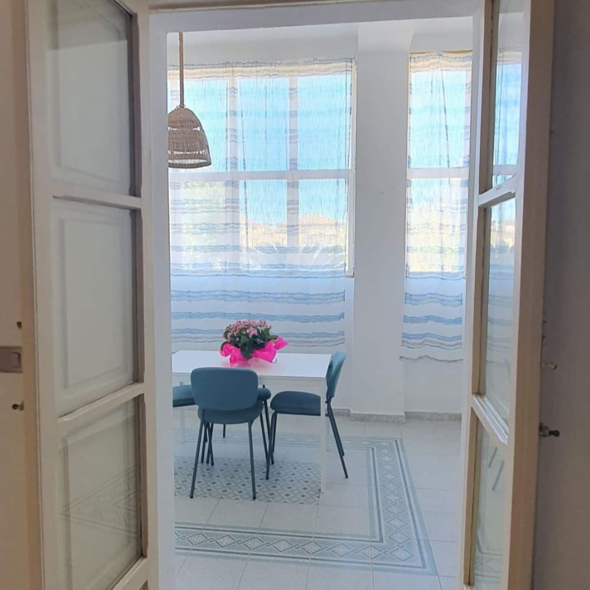 A light-filled dining area is visible through an open doorway, featuring a white table surrounded by four blue chairs. A floral arrangement with a pink ribbon sits at the center. Sheer curtains allow natural light to illuminate the space, while decorative tiles enhance the floor.